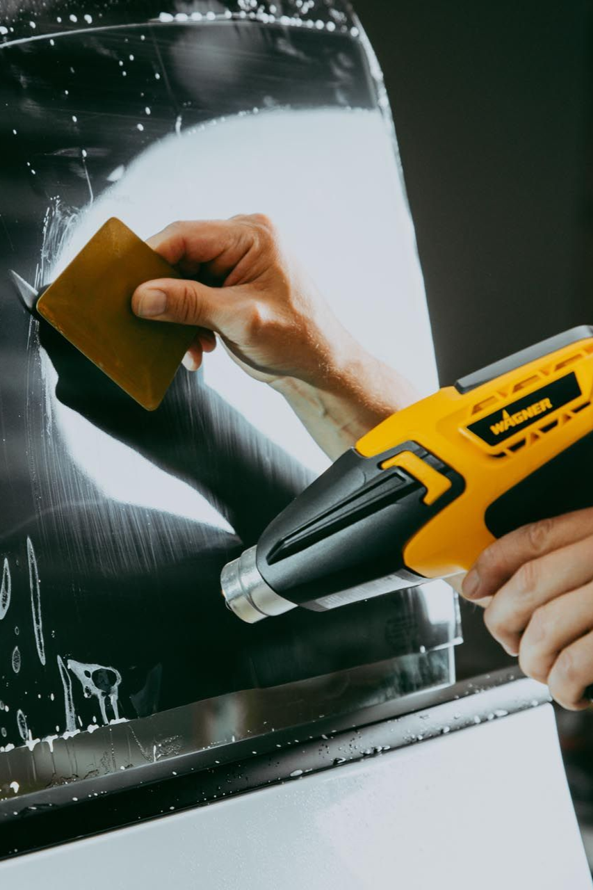 Person using a heat gun and a squeegee applying window film to a car window.