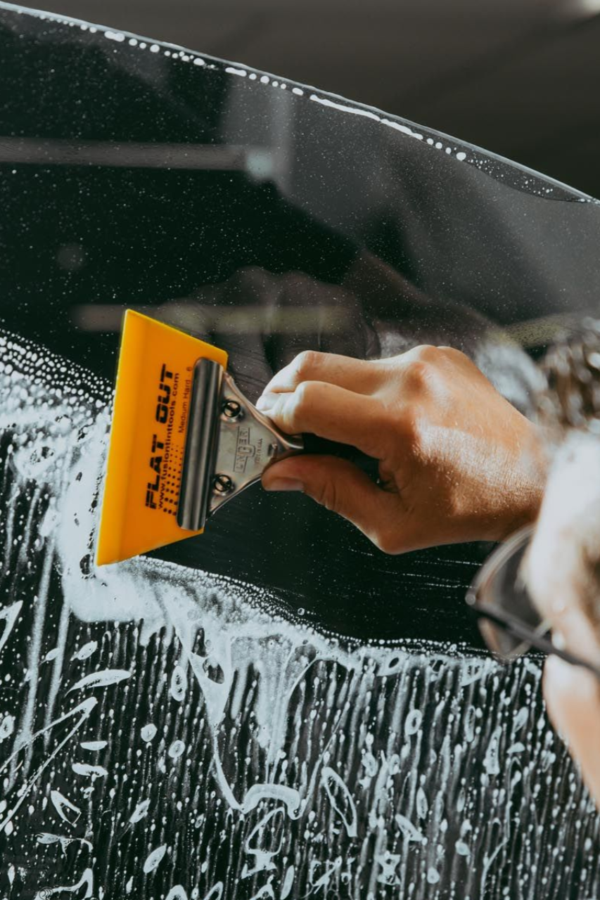 Person applying window tint with a yellow squeegee. Soap and film visible on car window.