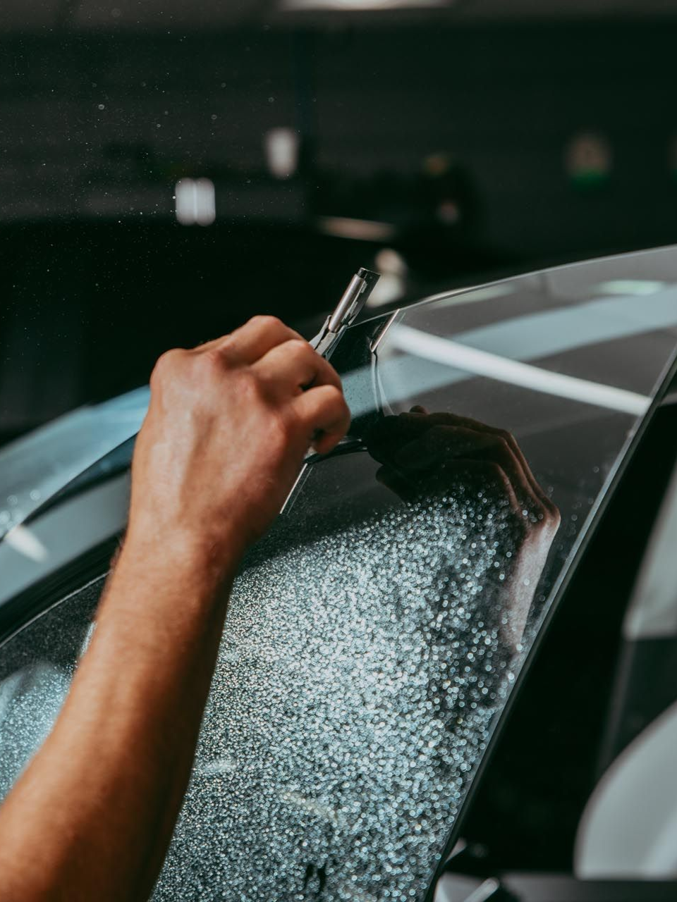 Person applying tint to a car window with a tool. Interior car setting, arm visible.