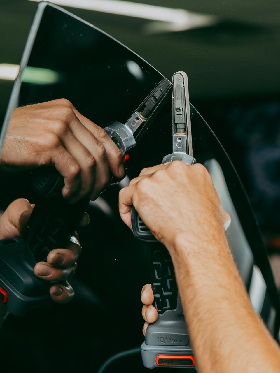 Hands using a tool to work on a car window. The tool has a red button.