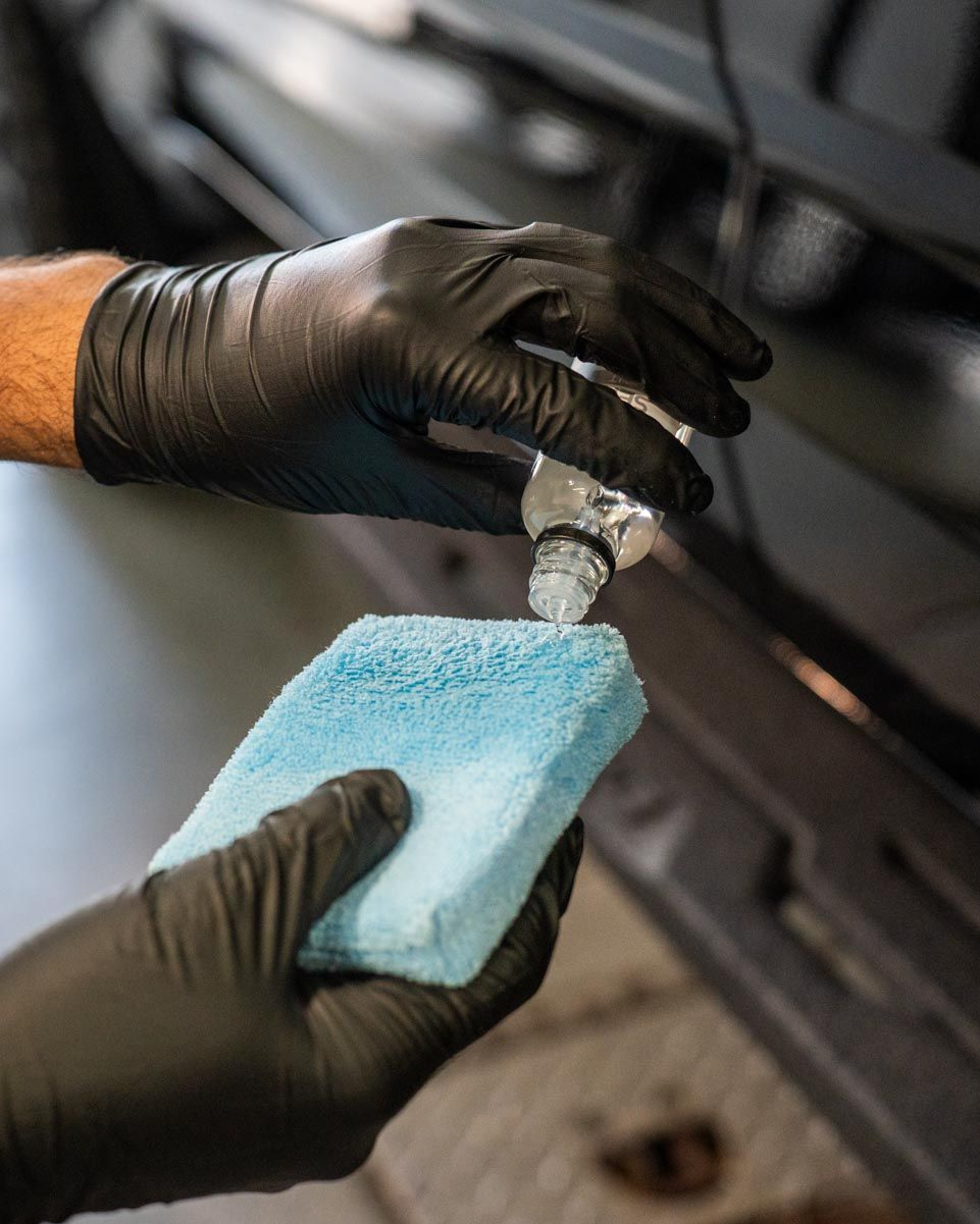 Gloved hands applying liquid from a bottle onto a blue sponge near a vehicle.