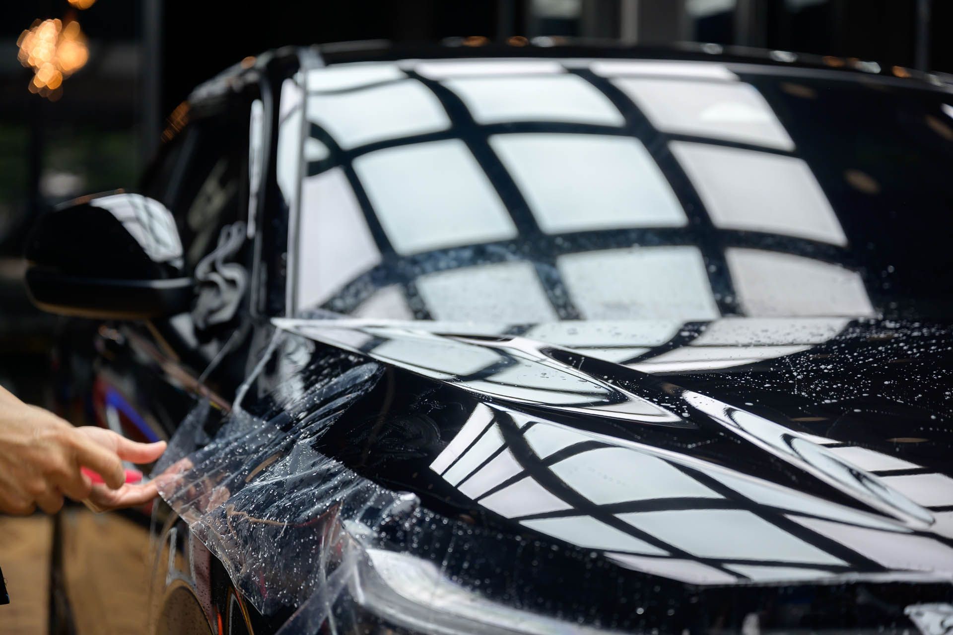 A person applying protective film to the hood of a black car. Reflective lights are above.