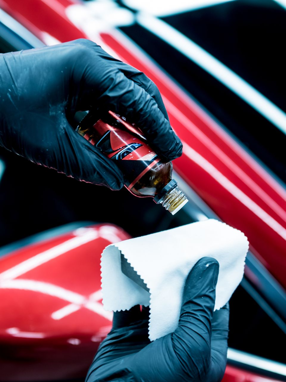 A gloved hand applying liquid from a bottle onto a white cloth, red car in background.