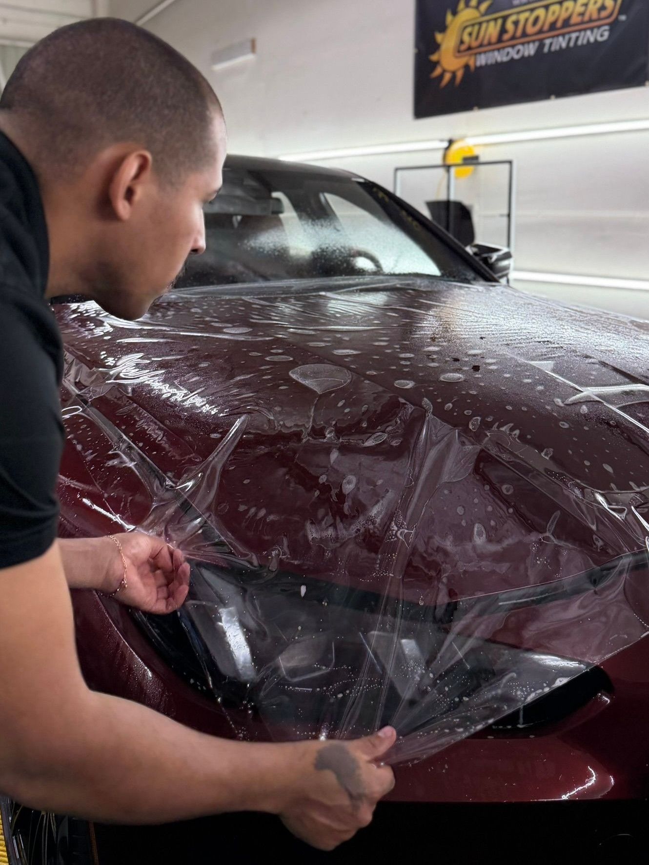 A professional applying a clear paint protection film to the hood of a dark red car in a workshop.