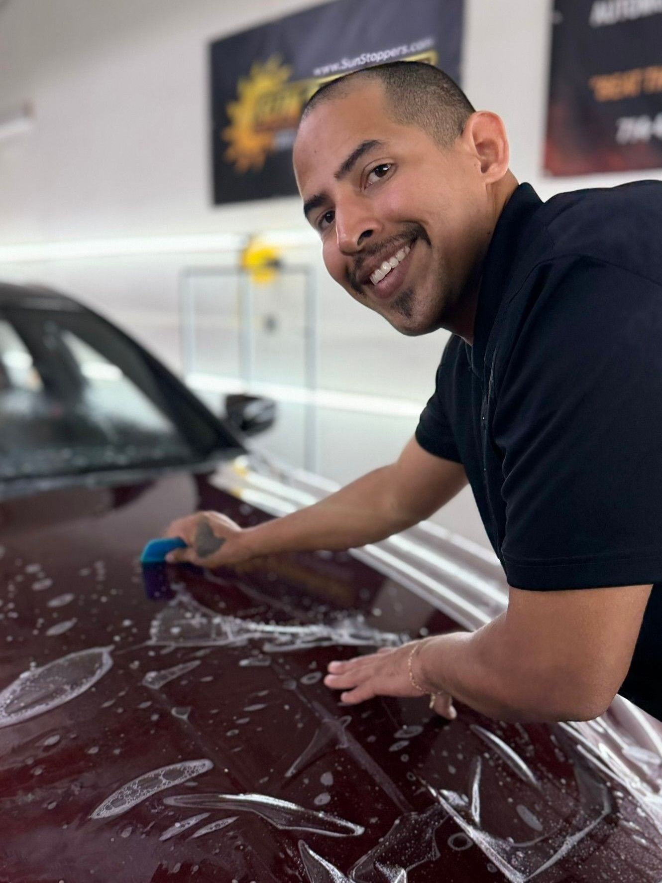 A technician smiling while applying a clear paint protection film to the maroon hood of a vehicle in a workshop.