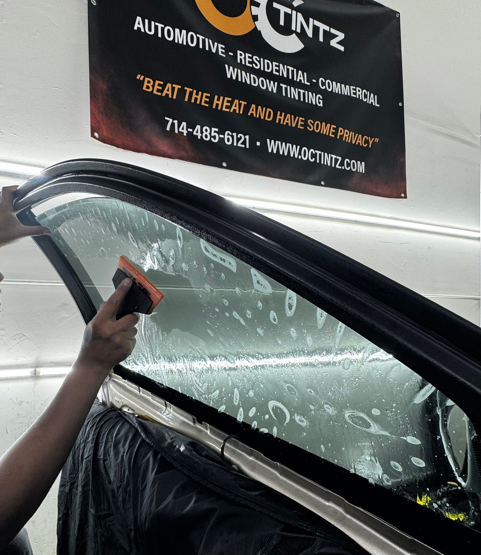 A person uses a squeegee to smooth window tinting onto a car window in a shop with a branded banner in the background.