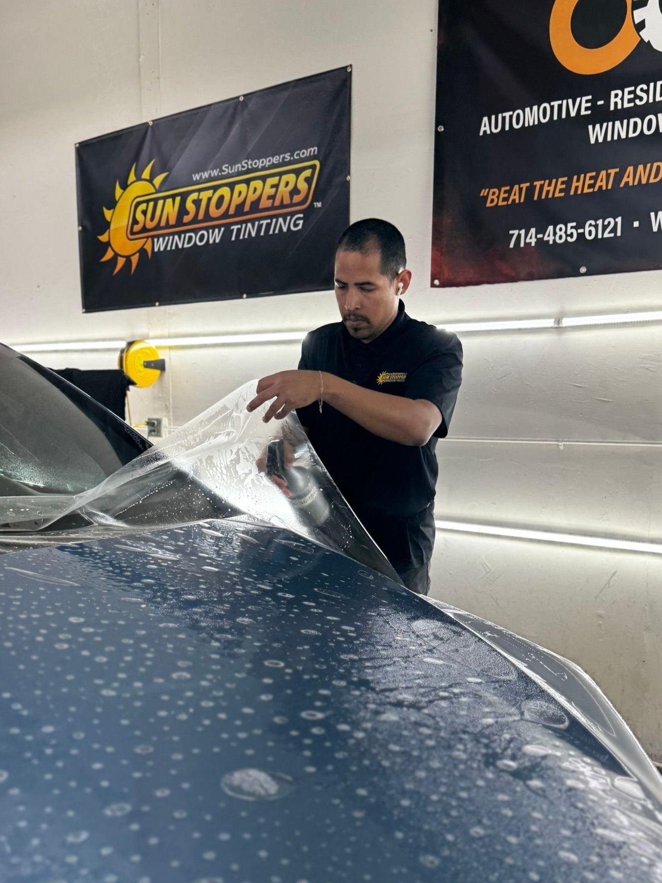 A technician in a black shirt applies a protective clear film to the hood of a blue car in a Sun Stoppers garage.