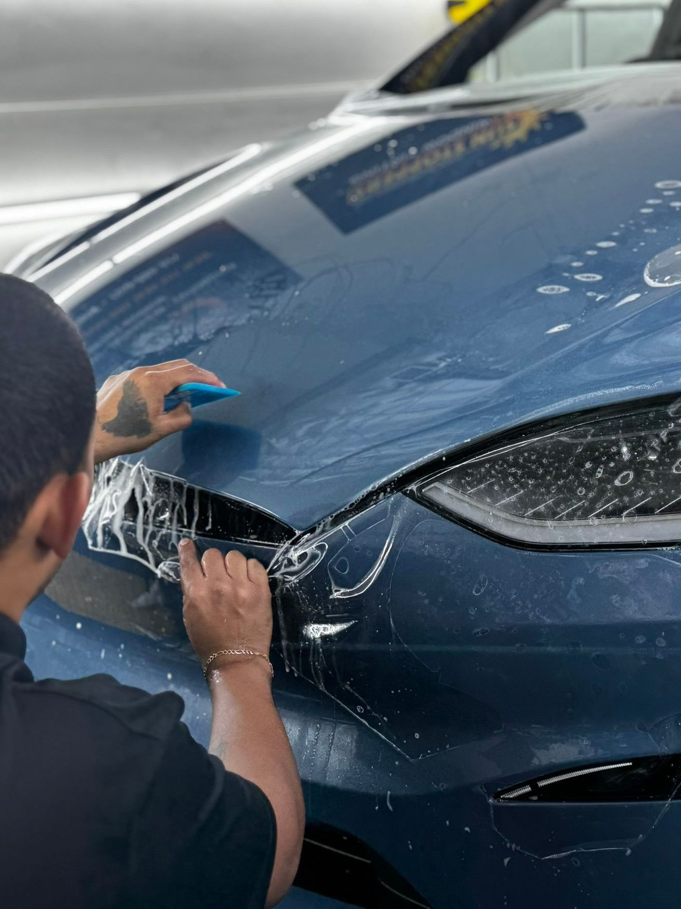 A technician uses a squeegee to apply a clear protective film to the blue hood of a vehicle.