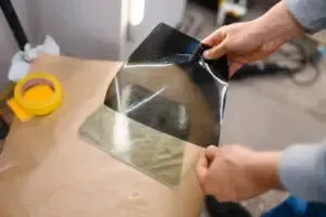 Hands holding tinted window film, showing varying opacity levels. Paper and tape in background.