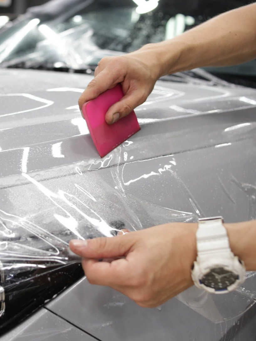 Person applying clear protective film to a car's hood with a pink squeegee, close-up.