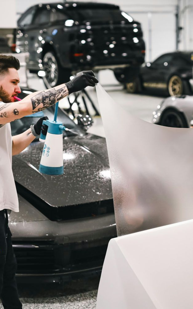 A man sprays a car with liquid while applying a wrap in a garage.