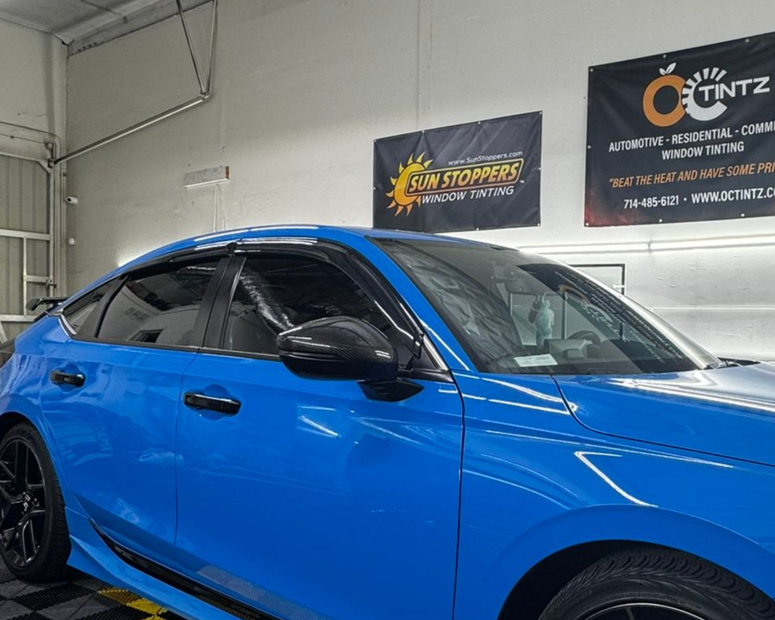 Person applying tinted film to a car window in a garage. Hands smooth out the dark film.