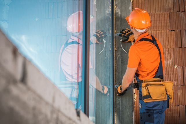 Construction worker in orange shirt and hard hat installing a window, exterior view.