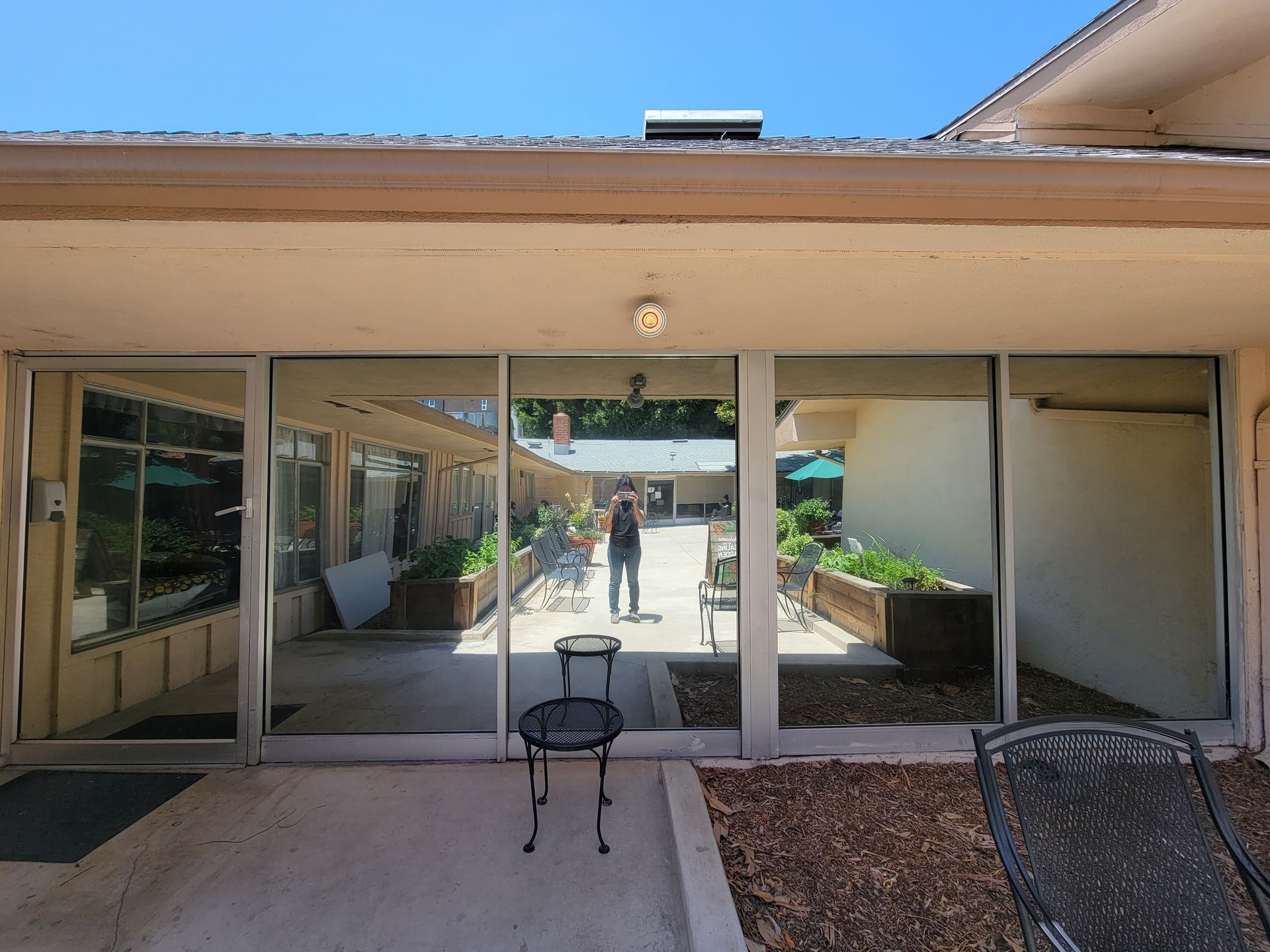 Glass doors reflecting a courtyard with a person, chairs, and potted plants. Beige building, sunny.