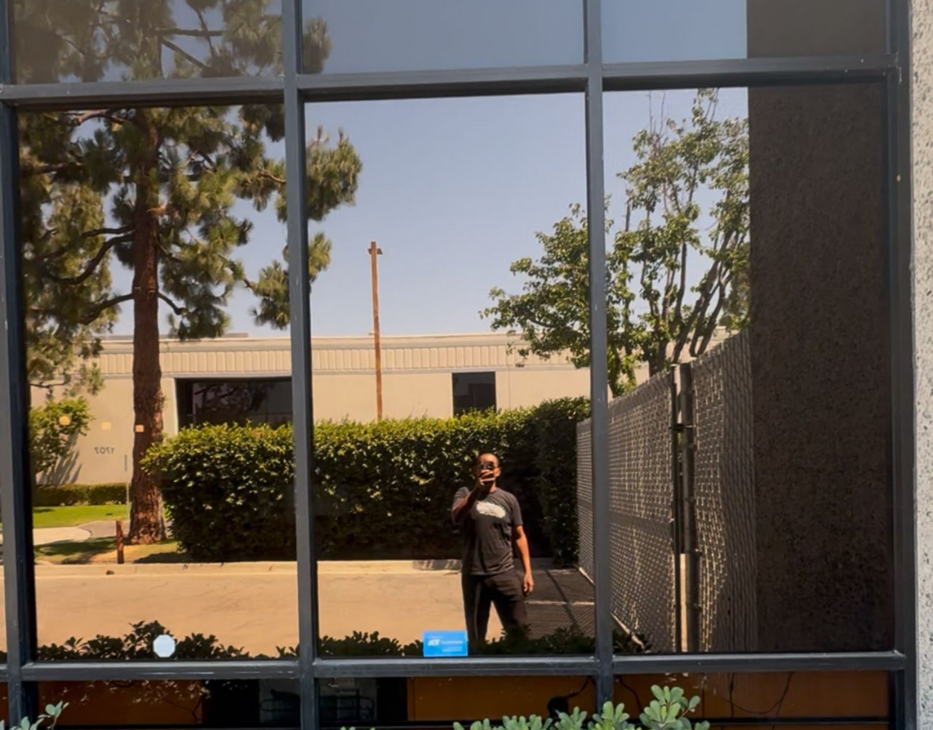 Man taking photo reflected in tinted window with outside view of buildings, trees, and blue sky.
