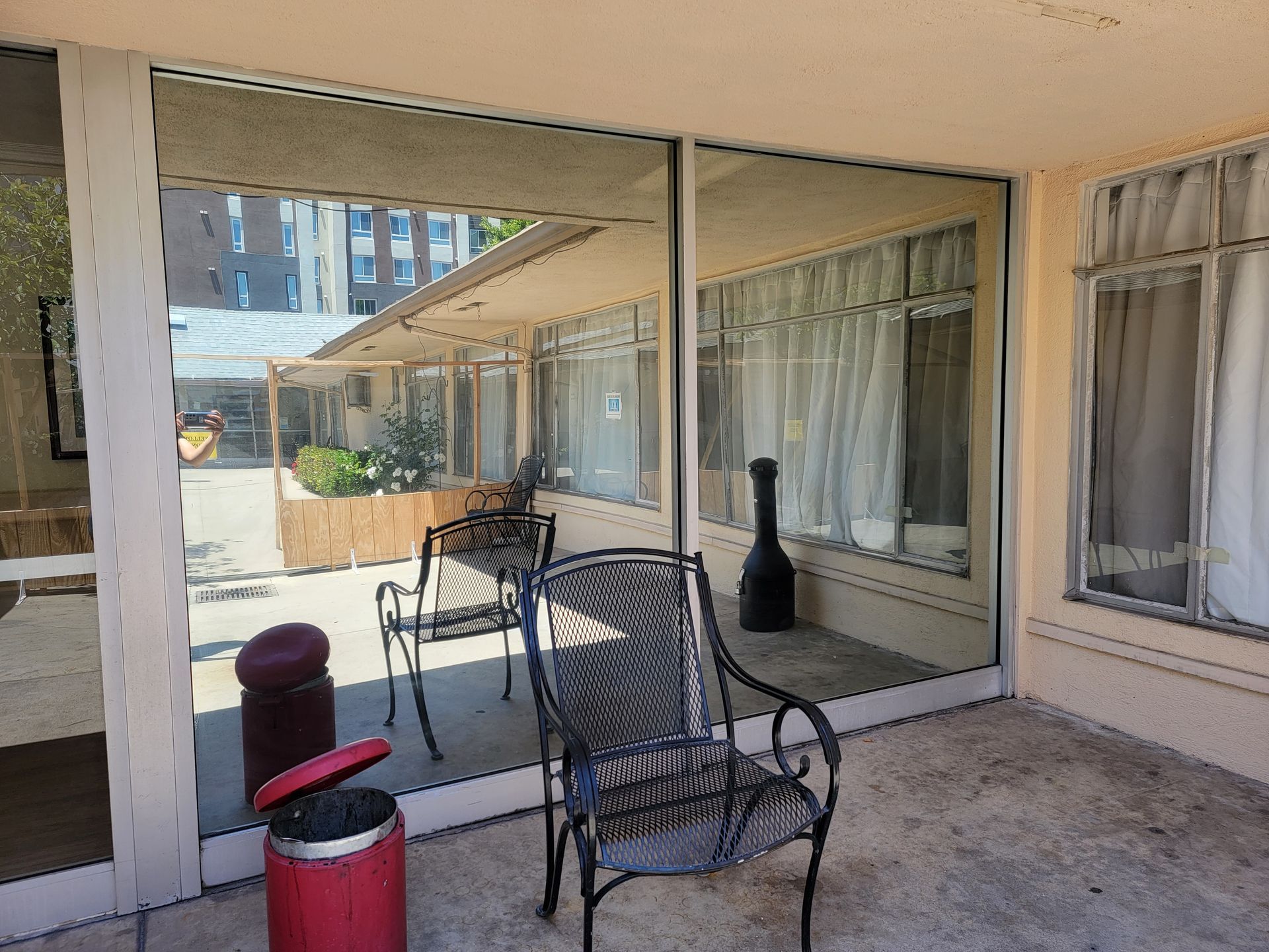 Patio with glass windows and doors reflecting the surroundings; metal chairs, a red trash can.