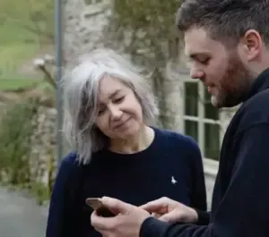 Woman and man looking at a phone outdoors. Woman with gray hair smiles. Man holds phone. Stone building in background.