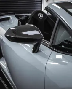 Close-up of a light-colored Lamborghini's side mirror, reflecting a carbon fiber finish, with black interior in background.