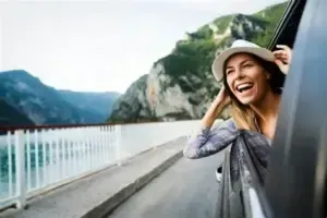 Woman with a sun hat laughs from a car window, scenic background of mountains and water.