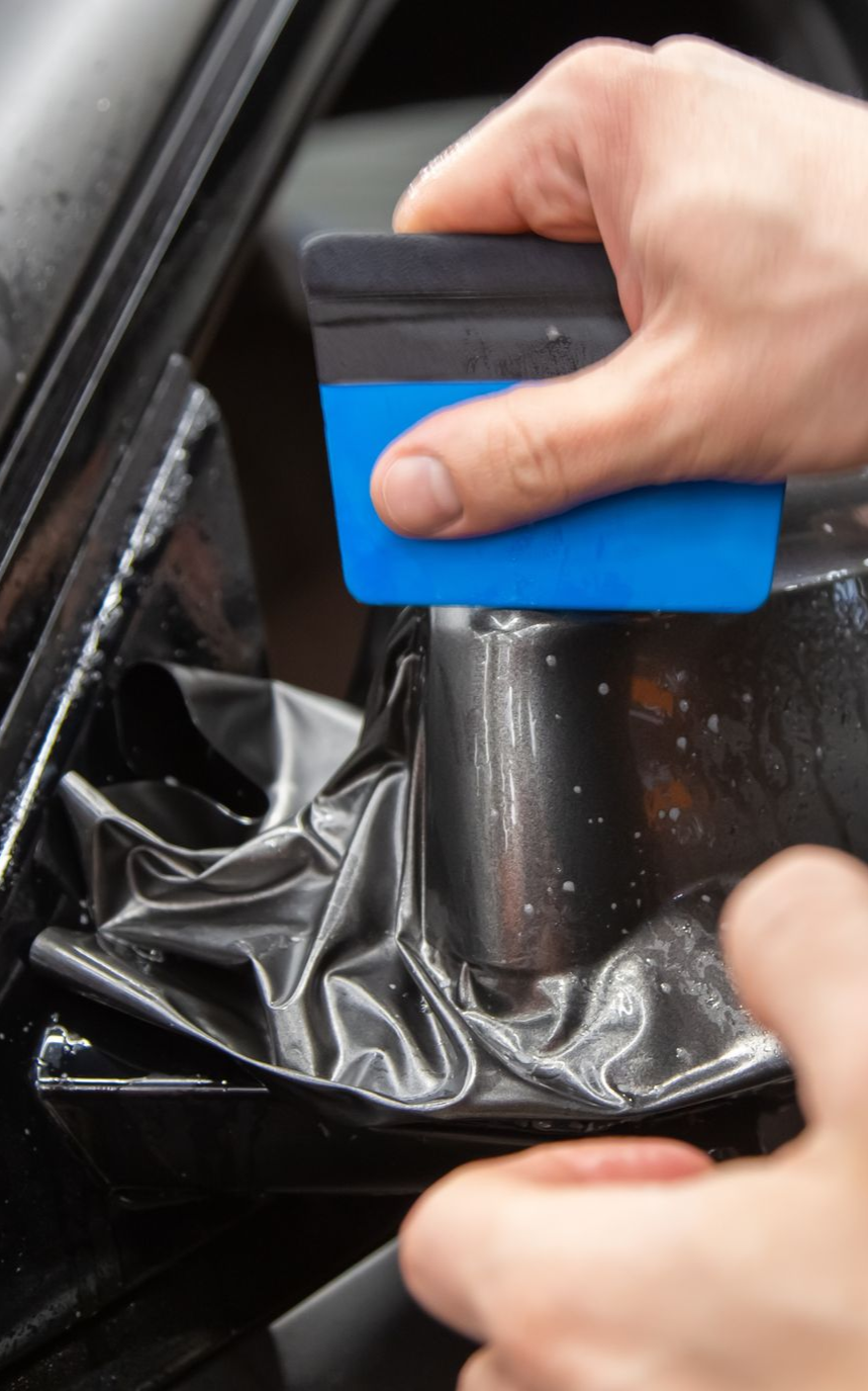 Person using a blue squeegee to apply black window tint to a car window.