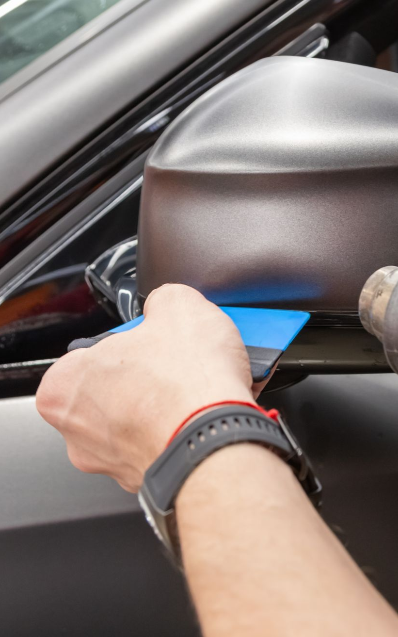 Person using a squeegee to apply material to a car's side mirror.