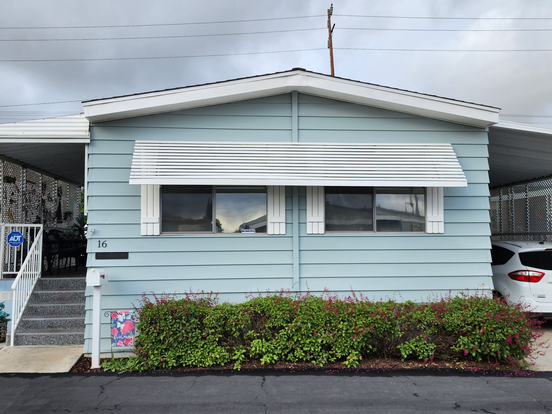 Light blue mobile home with awning, bushes, and parked car.