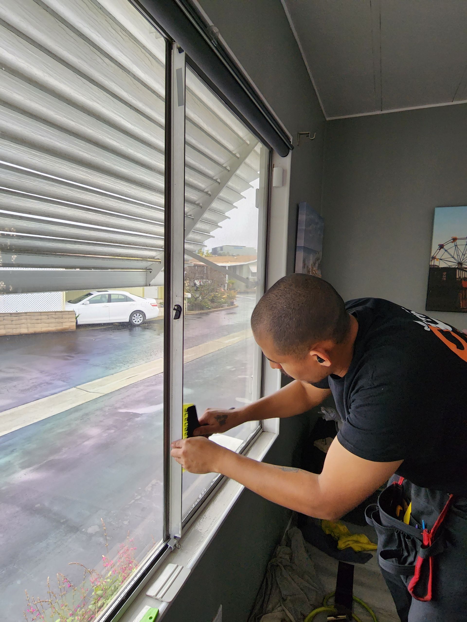 A man installing window tinting on a glass window in a room. The window overlooks a parking lot.