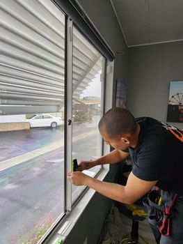 A man applying window tint in a room. He's wearing black, using a yellow tool, indoors with outside view.