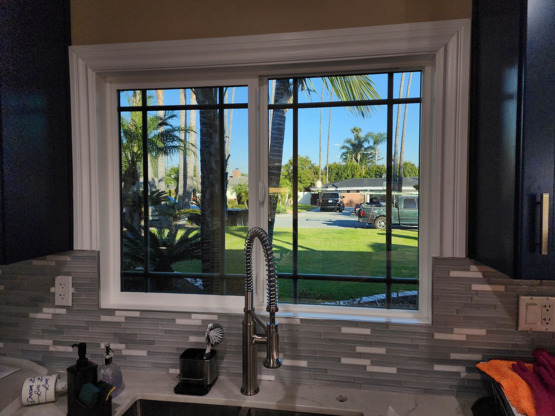 Kitchen window with a view of a sunny yard. Black grid, white trim, and a stainless steel faucet.