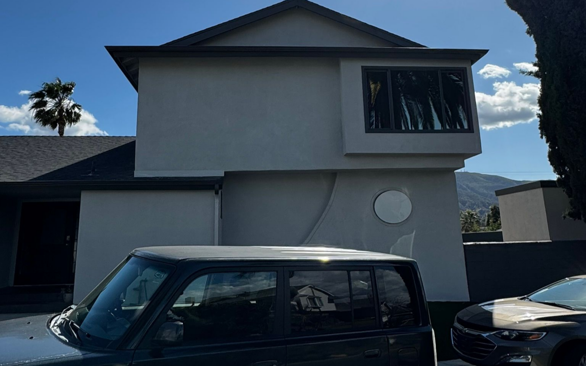 A dark green Scion xB parked in front of a light gray house with black roof and a window.