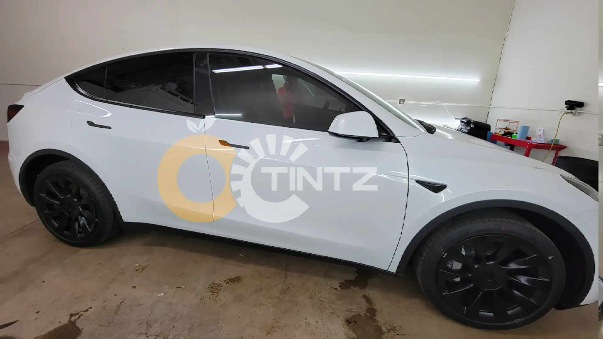White Tesla Model Y with black wheels in a shop with tinting logo.