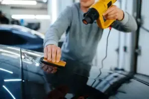 Man using a heat gun and squeegee on a car's dark surface, presumably applying tint in a workshop.