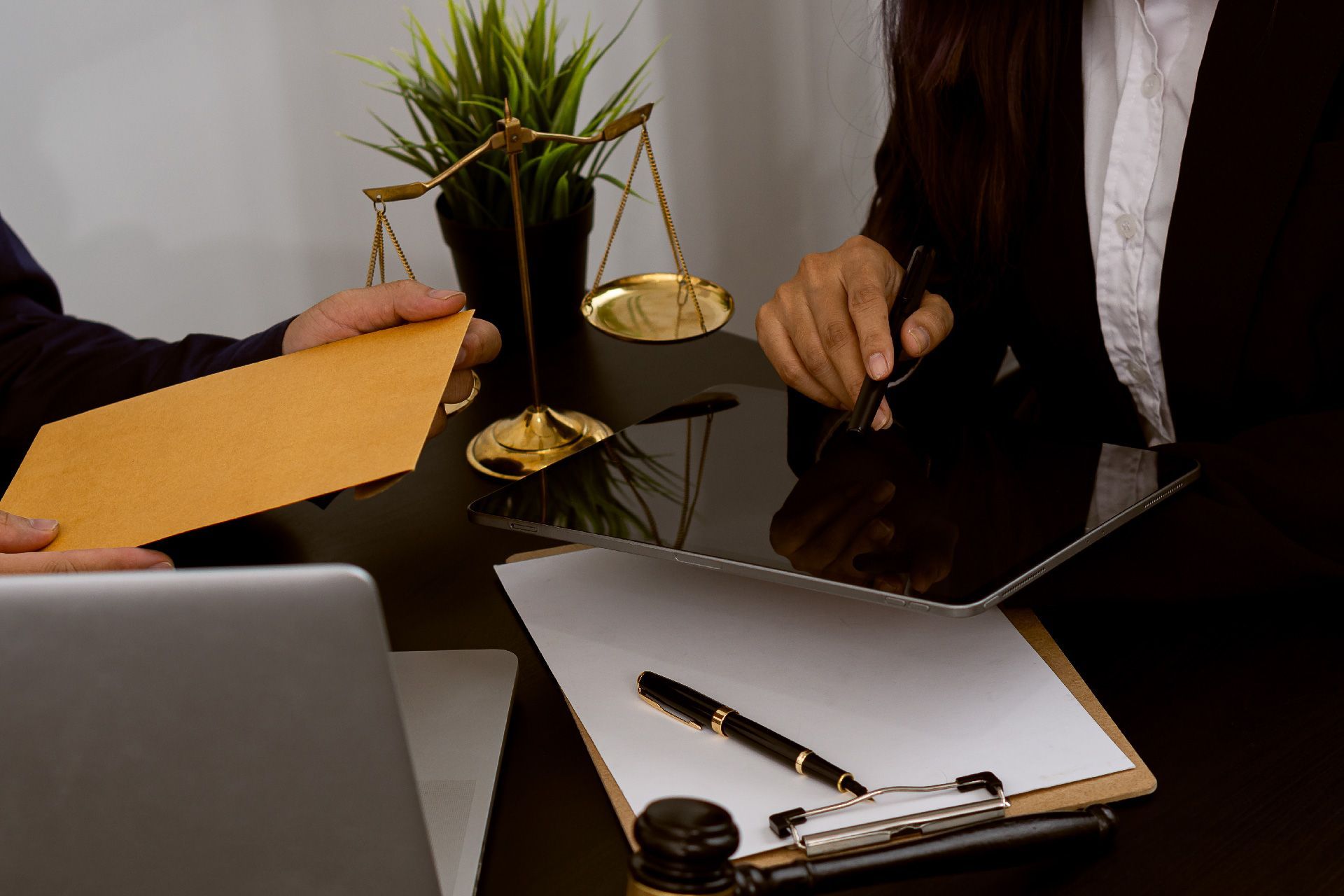 Persona entregando documento a otra, con balanza de justicia, computadora portátil y bolígrafo en el escritorio.