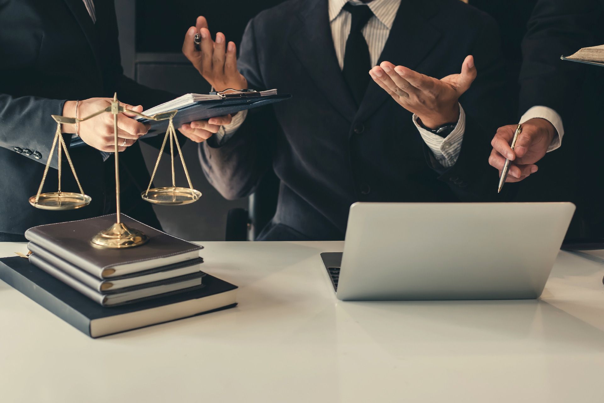 Tres abogados de traje discutiendo un caso en un escritorio con balanzas de justicia, libros y una computadora portátil.
