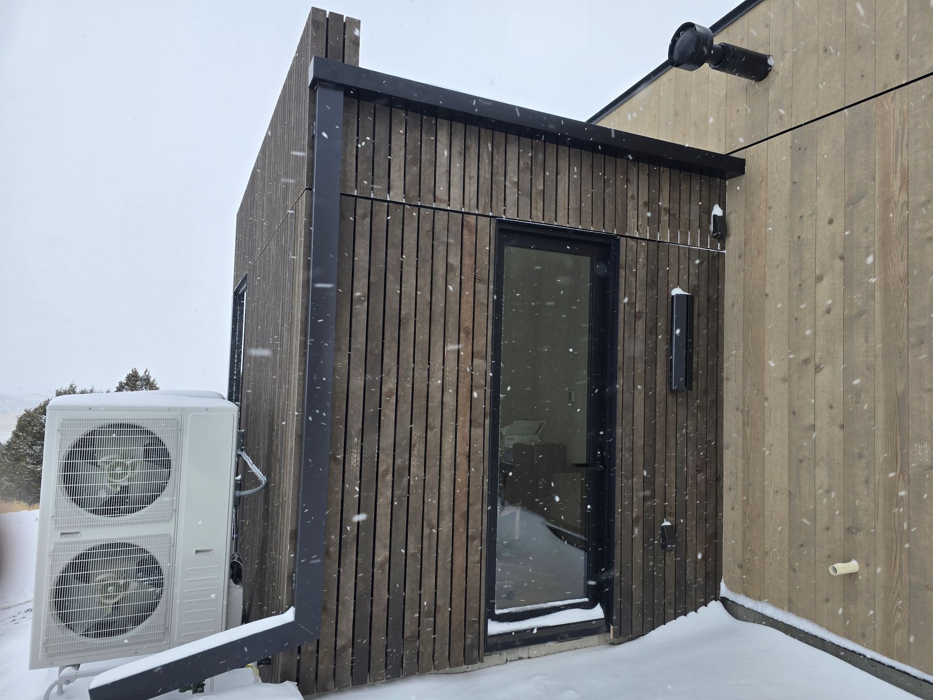 A small, vertical-slat wooden structure with a black door and adjacent HVAC unit sits on a snow-covered deck.