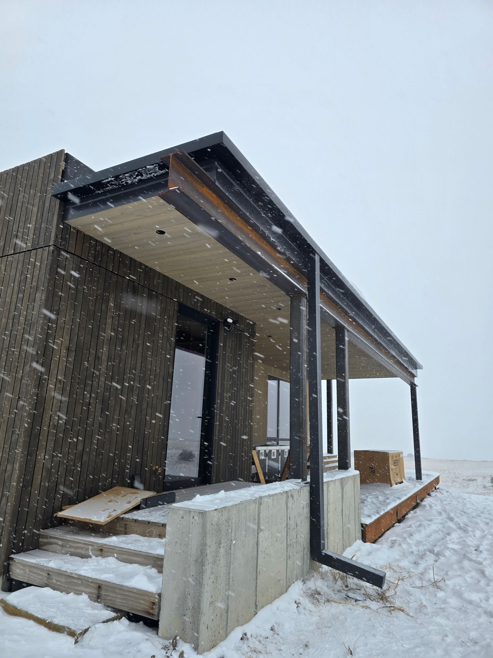 A modern house with textured brown siding and a concrete porch with stairs, set in a snowy, overcast landscape.