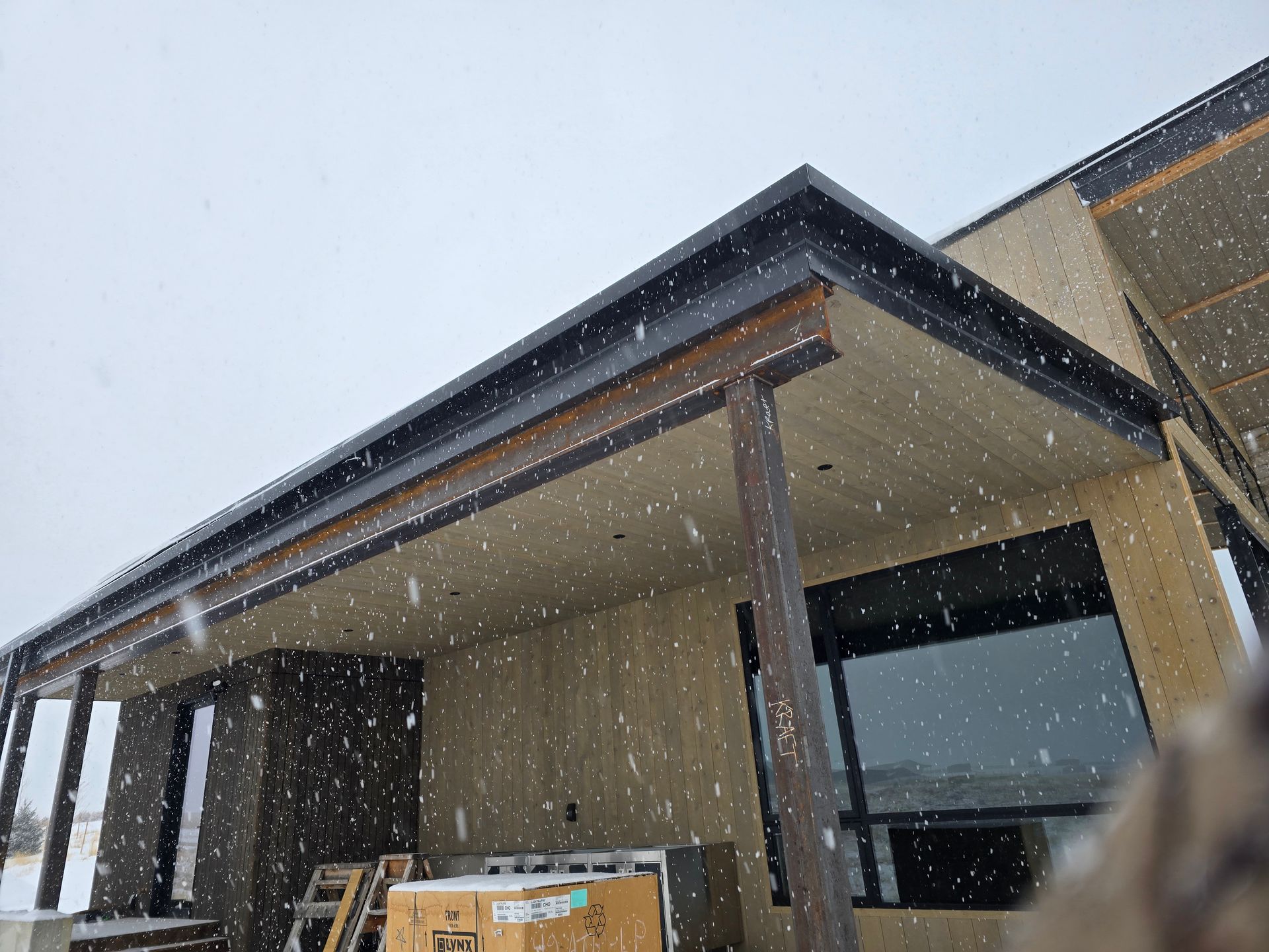 A modern home patio with a dark metal beam structure, wood-paneled ceiling, and large windows during a snowfall.