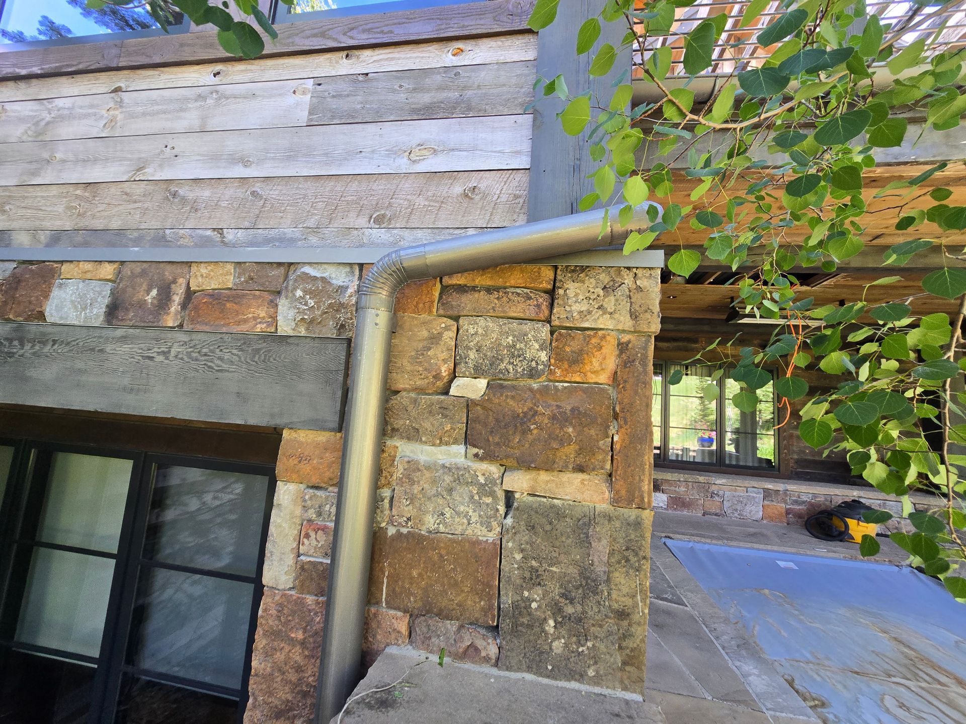 A metal downspout runs along a stone wall beside a window, with wooden siding above and a patio visible in the distance.