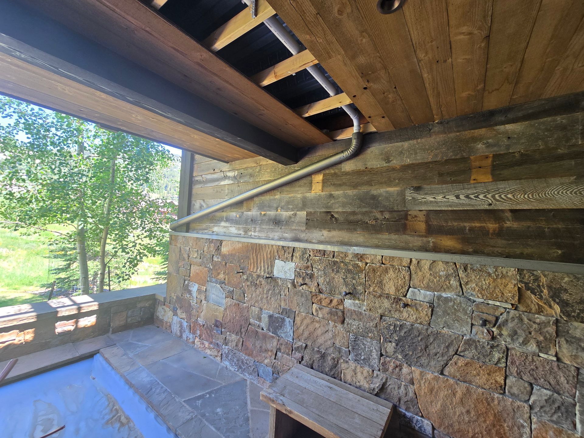 A rustic stone wall supports a wooden beam ceiling with a metal drainage pipe running diagonally across the stones.