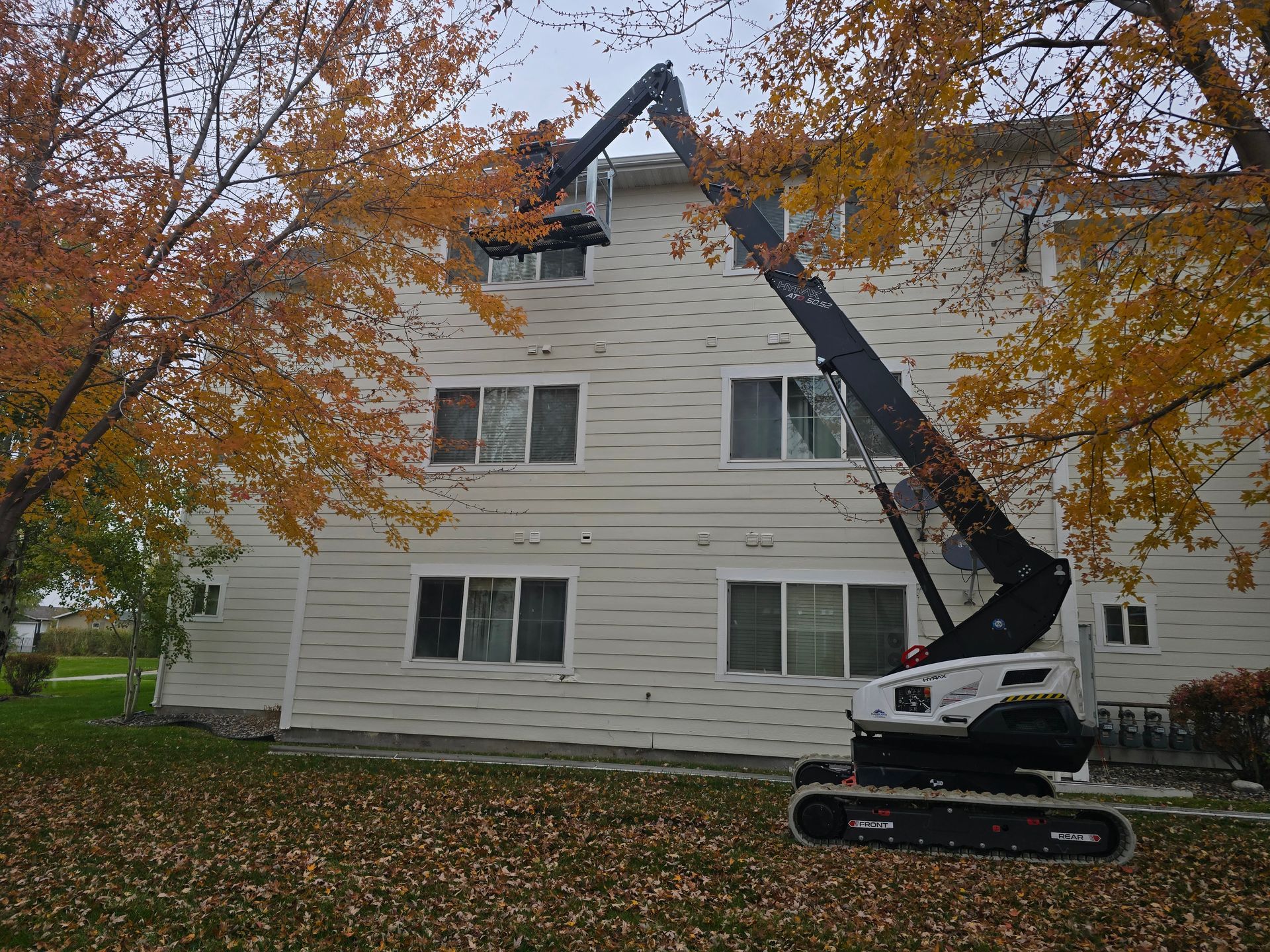 A white tracked lift reaching up to the gutter of a three-story beige apartment building during autumn.