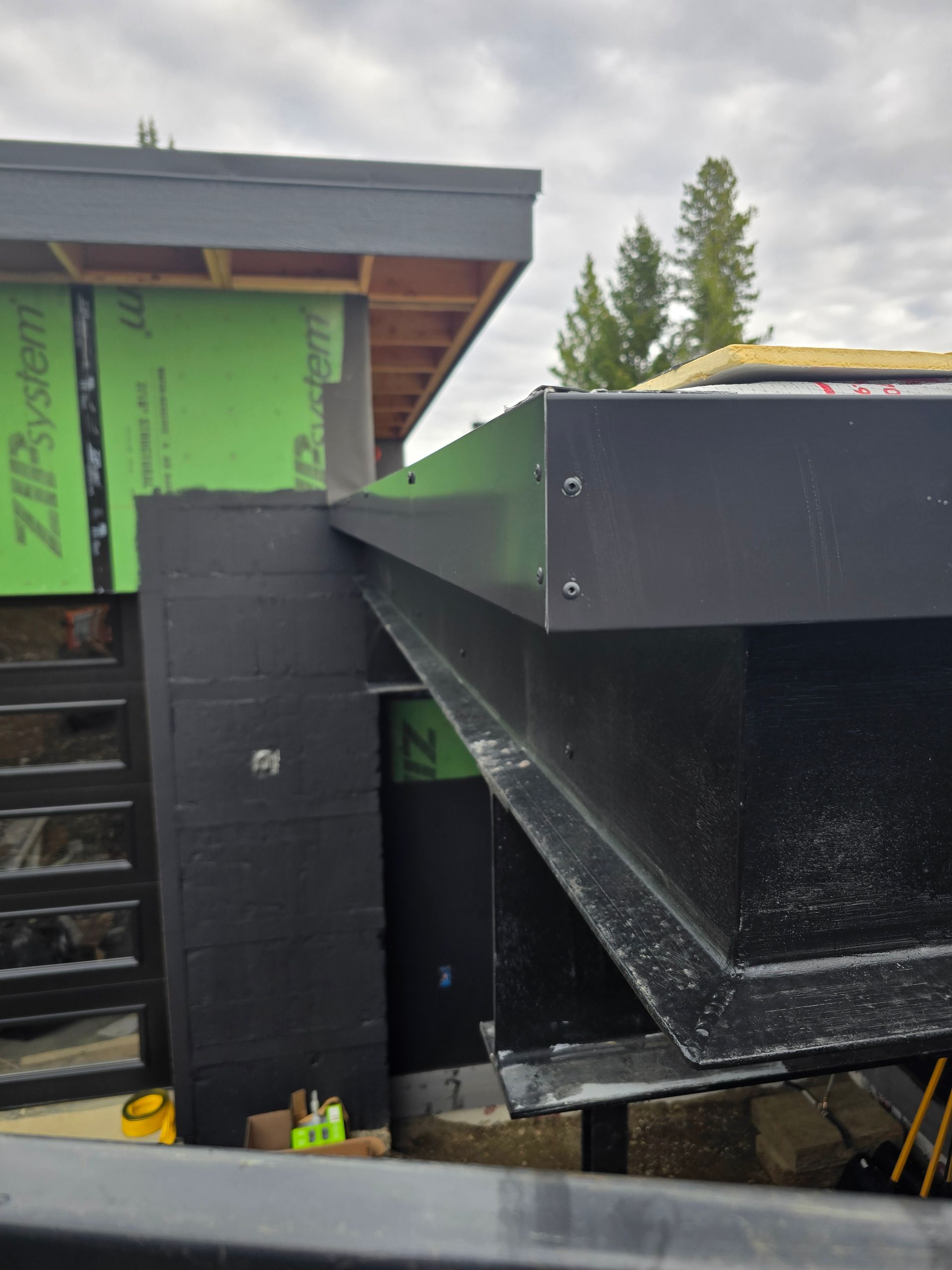 Construction view of a black metal beam, roof edge, and green ZIP System sheathing on an unfinished modern building.