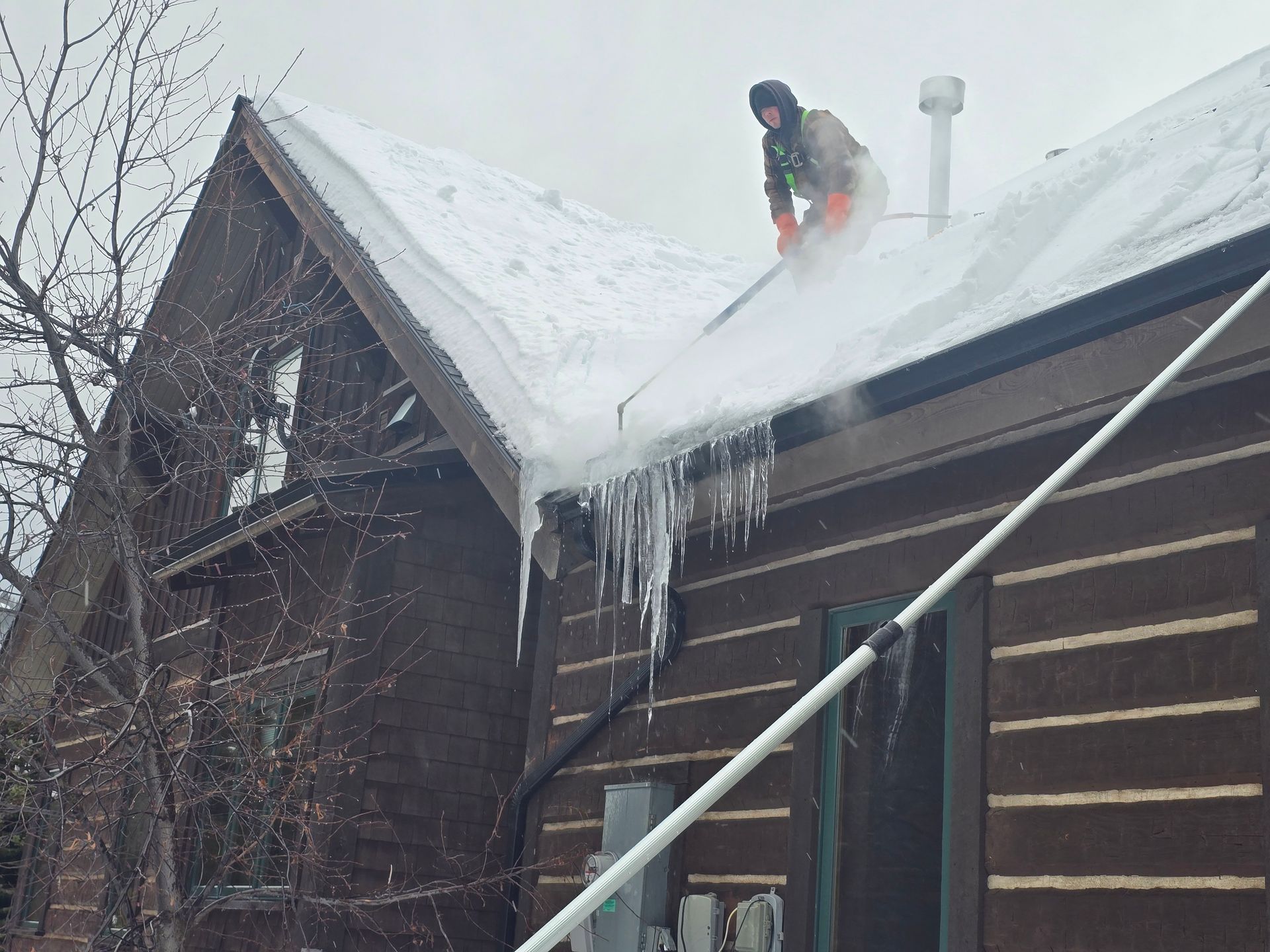 A person on a snowy roof uses a long pole to clear heavy snow and icicles from the eaves of a brown wooden cabin.