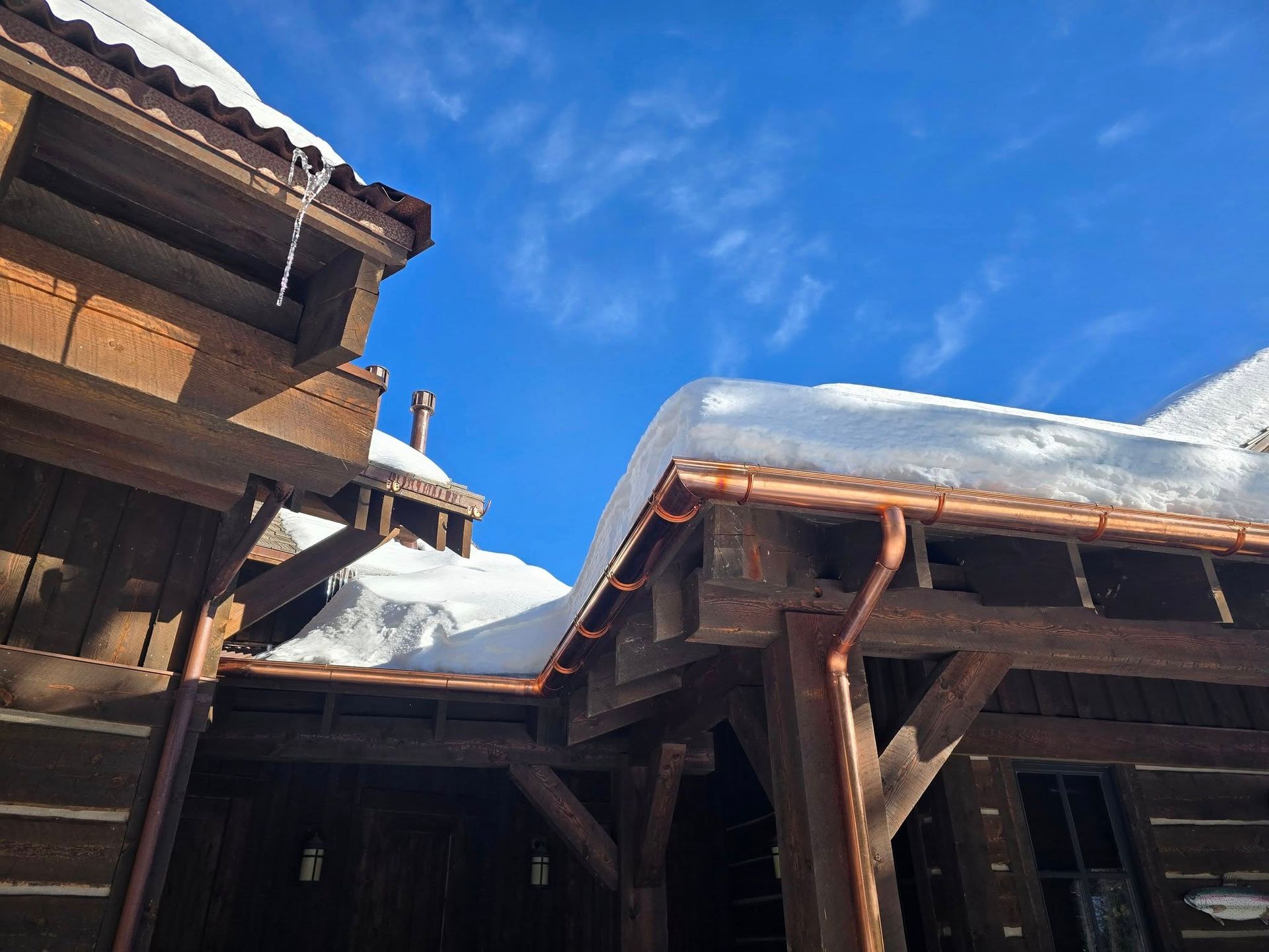 Low-angle view of a dark wooden rustic house with copper gutters, covered in heavy snow against a bright blue sky.
