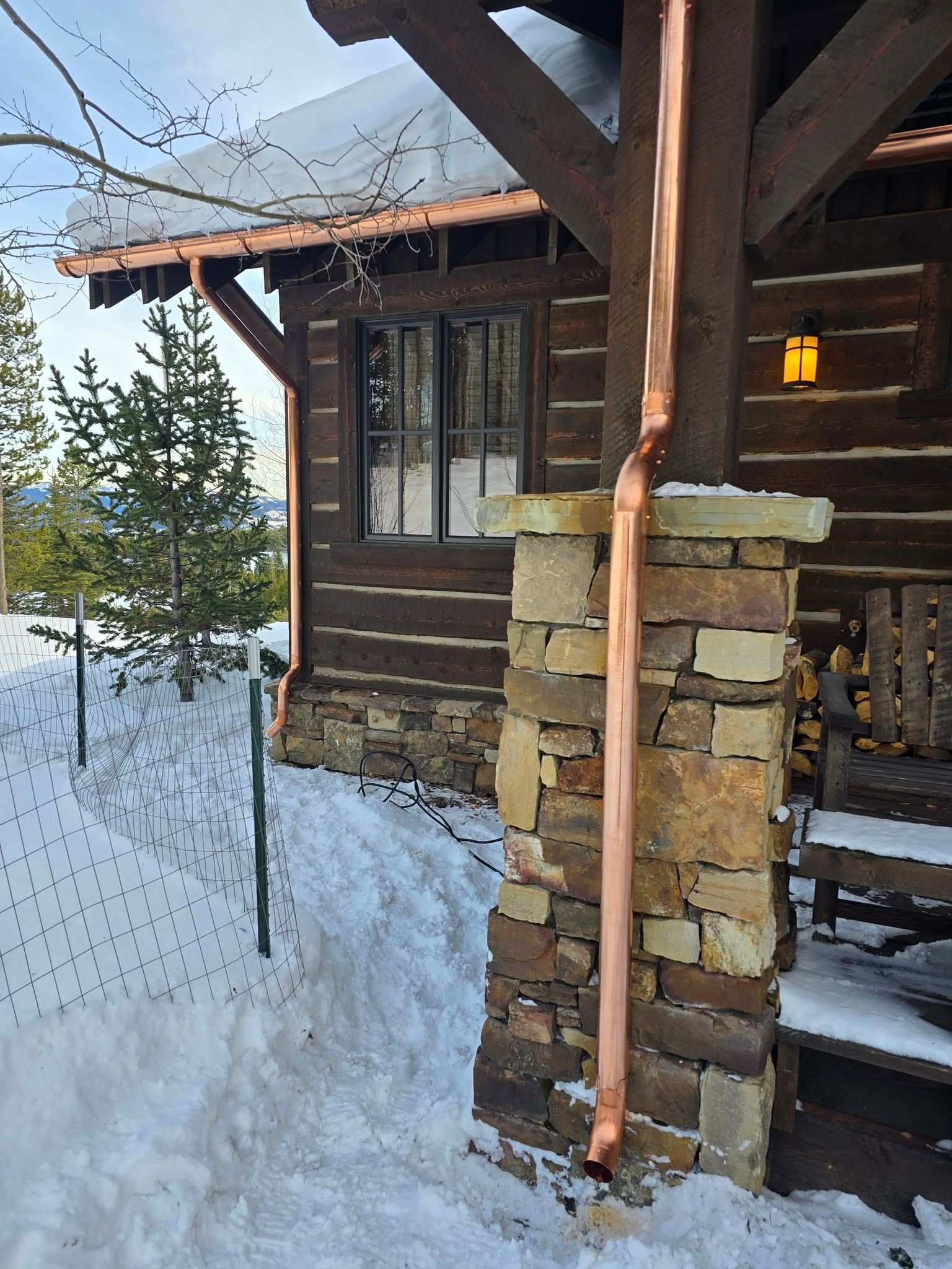 A copper downspout runs down a stone pillar beside a rustic log cabin covered in snow.