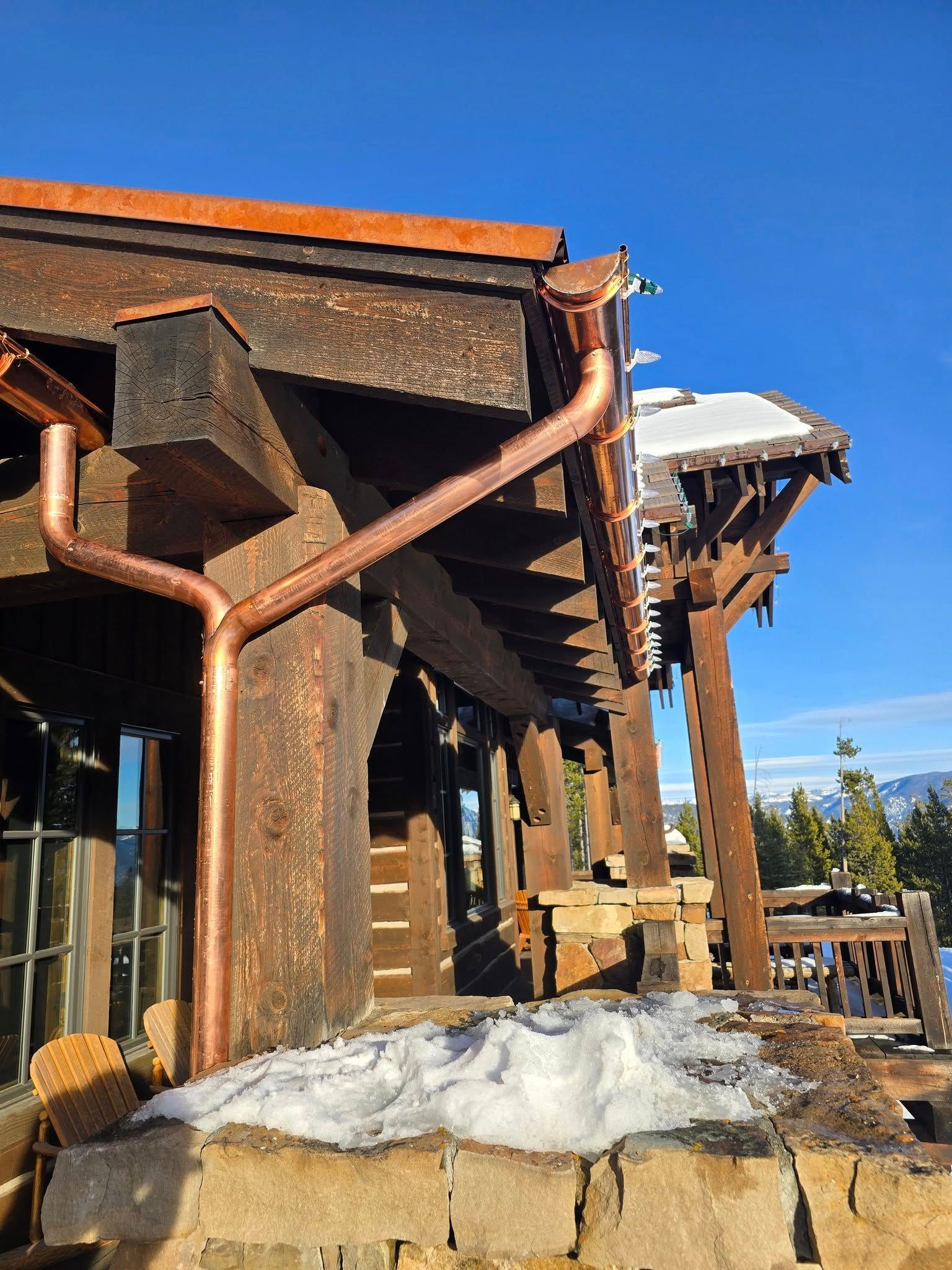 Copper gutters and downspouts on a rustic wooden building with snow on the roof and stone base against a blue sky.