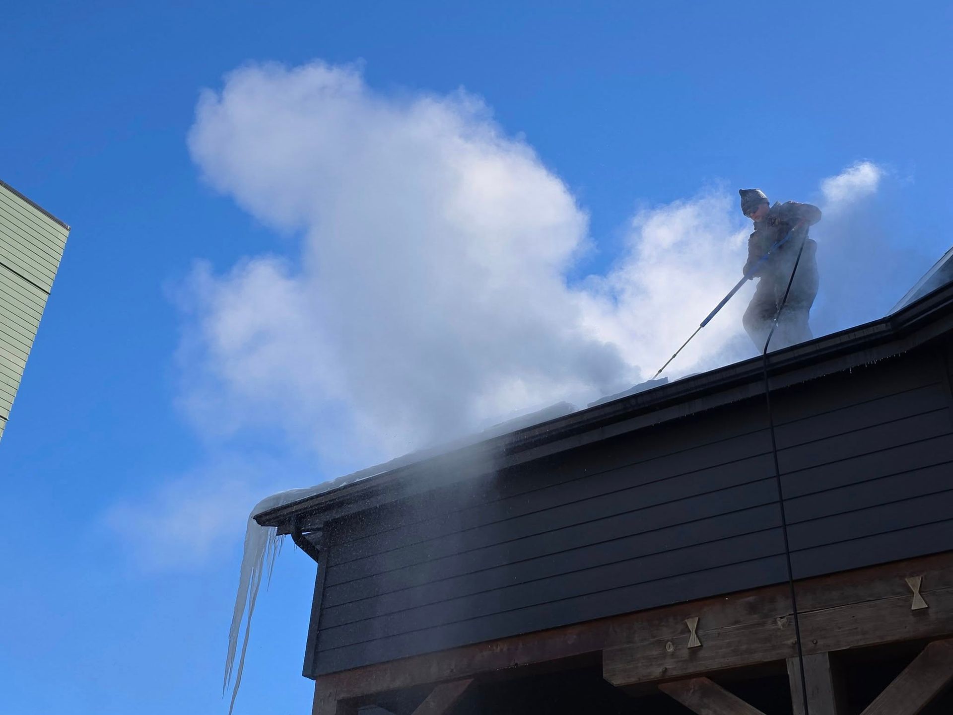A person on a ladder uses a long-handled tool to clear icicles from the edge of a dark wooden building roof.