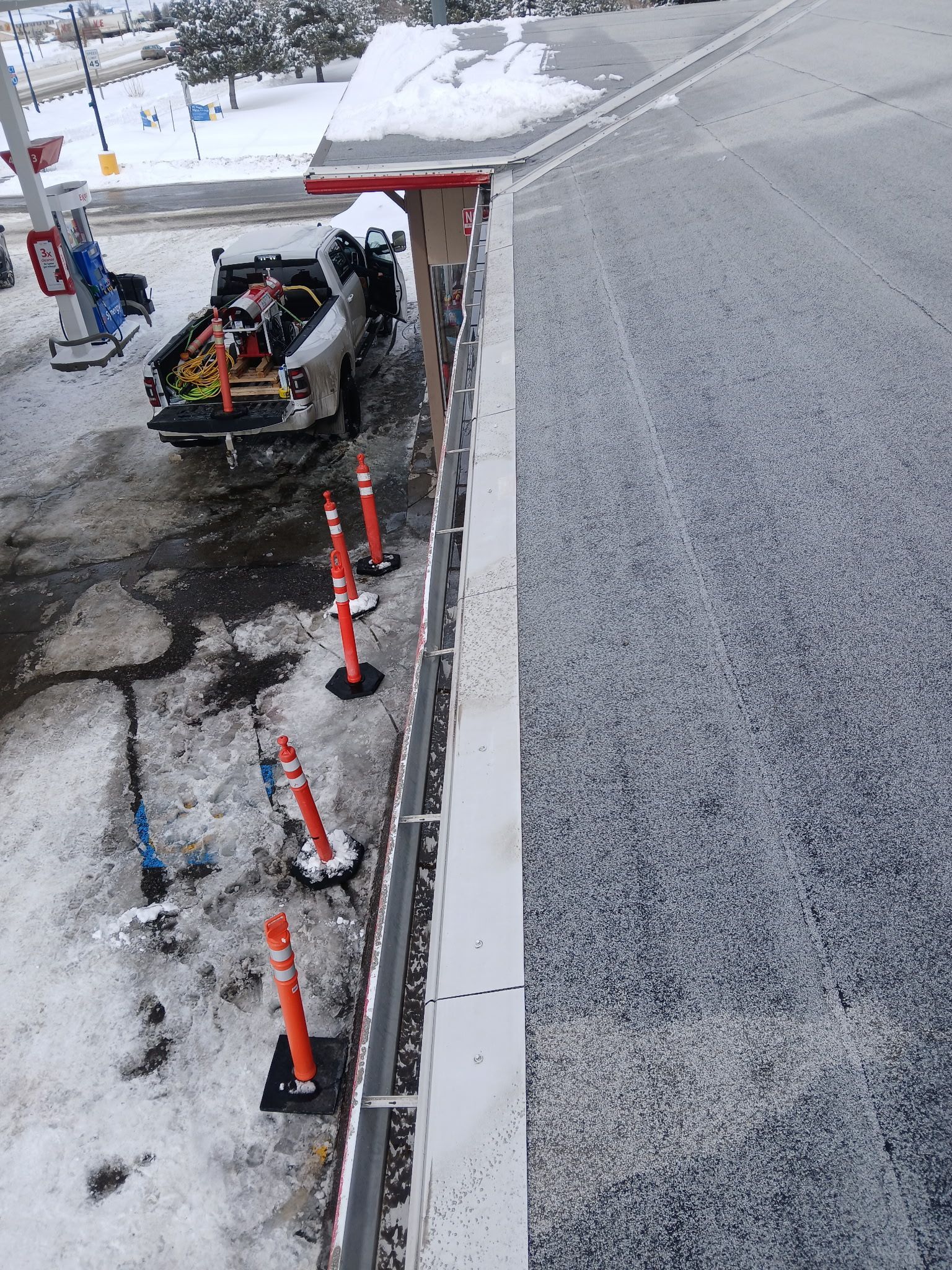 An elevated view shows a snow-dusted roof edge, a service truck with equipment, and orange safety cones on snowy ground.