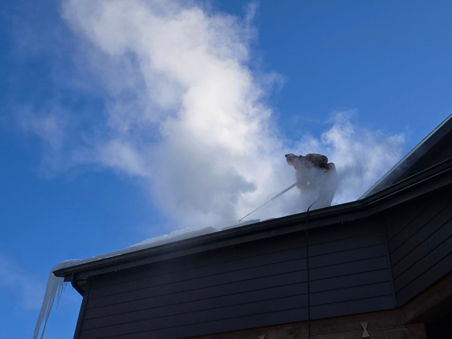 A person using a tool to clear snow and ice from a residential rooftop against a bright blue, cloudy sky.