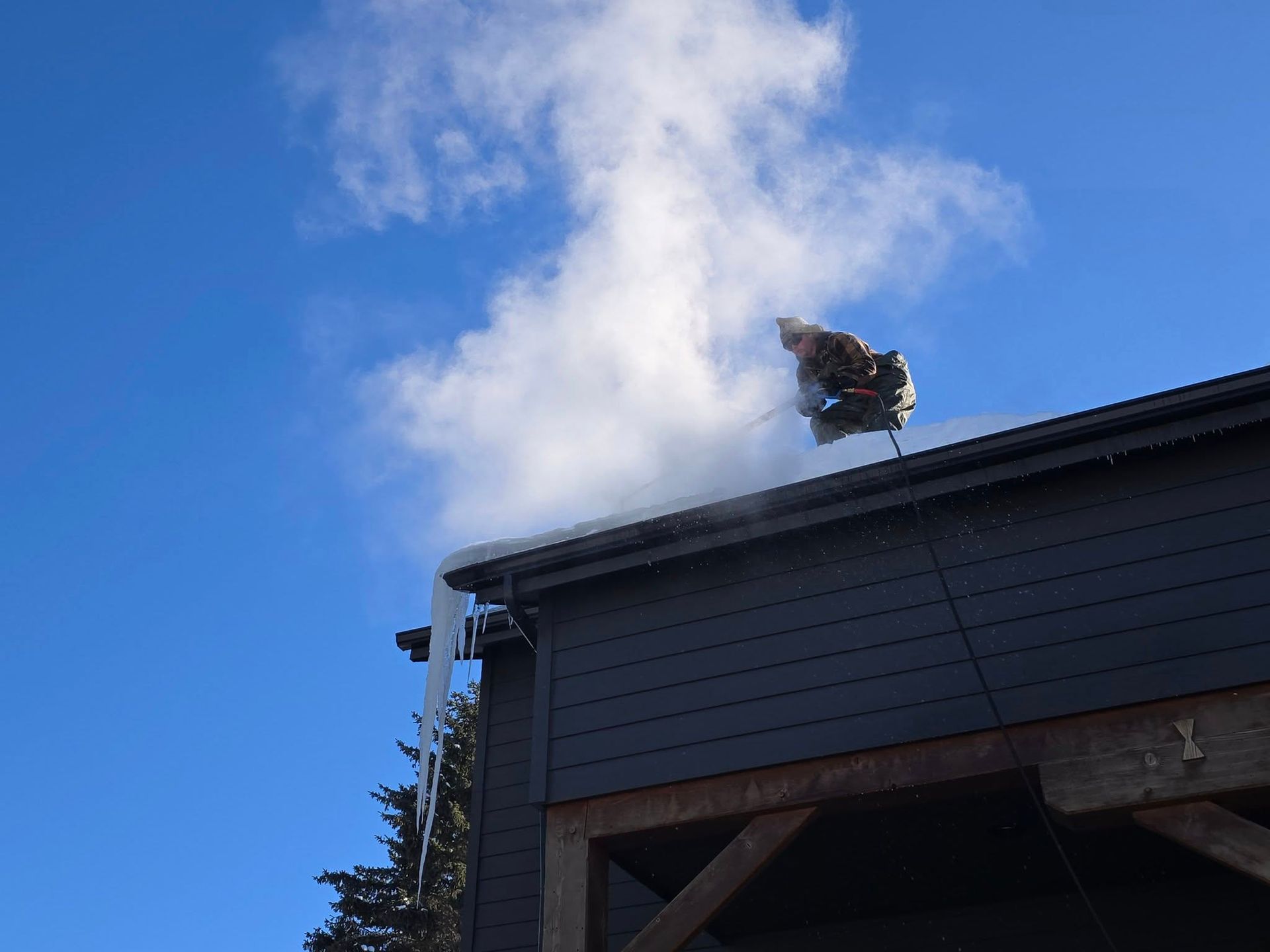 A dark-sided building corner under a clear blue sky, featuring a small steam plume and a hanging icicle on the roof edge.