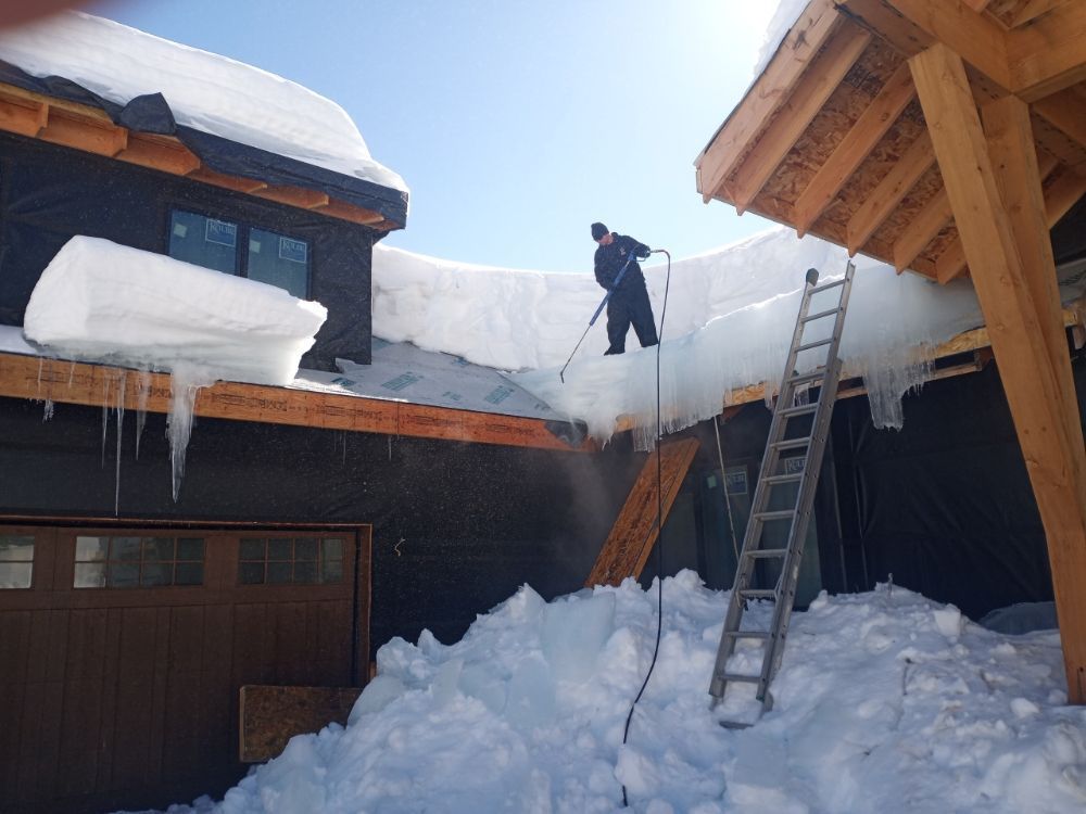 A person stands on a snowy roof clearing ice with a tool, near a ladder and a large pile of snow by a house.